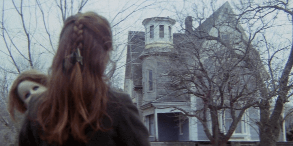 A still from the film The House by the Cemetery. A young girl carrying a doll looks at a house.