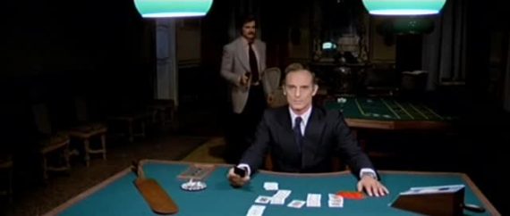 A still from the film Gang War in Milan. A man sits at a casino table, with another man approaching him from behind.